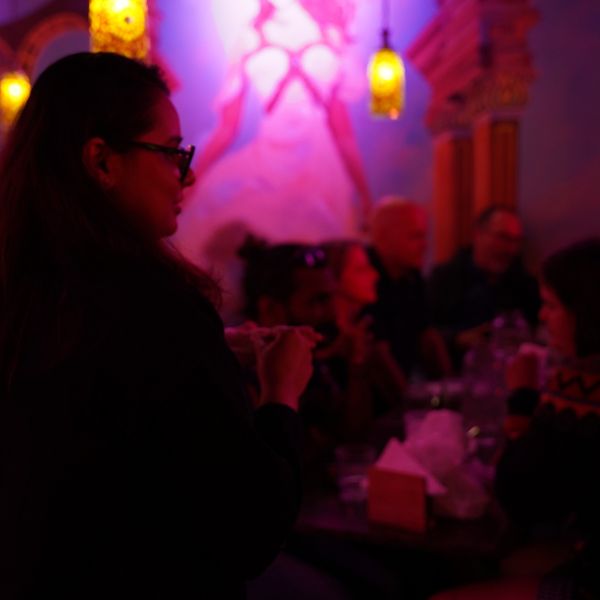 A woman in glasses enjoys a lively dinner with friends at a cozy table in a bustling Indian restaurant on Gerrard Street.

